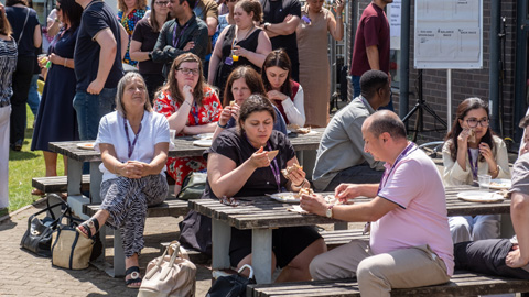 People gathered outdoors around picnic tables, eating, drinking, and talking near a brick building with posters